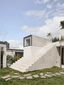 Modern, minimalist staircase constructed of concrete, featuring a single window and surrounded by a green landscape with ston