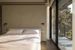 A modern minimalist bedroom with a wooden floor, white linens, and a large window overlooking greenery.