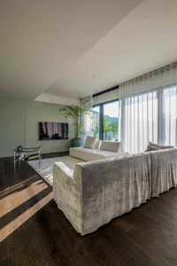 A modern living room features grey couches on dark wood flooring with large glass windows.