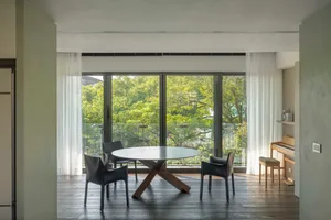 Modern living room with wooden floor and black furniture, white curtains, and large windows showing green trees outside.
