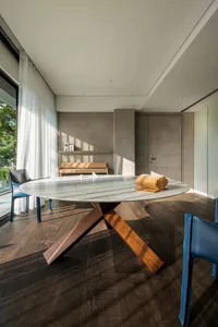 A modern dining room featuring a round marble table with a wooden base, blue chairs, and sunlight streaming through the windo