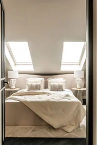 Modern, minimalist bedroom with white bedding, pillows, and a light-colored fabric headboard, featuring skylights and a cozy 