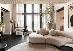 Modern living room featuring a curved, beige fabric sofa, dark wood flooring, and large windows with white curtains.