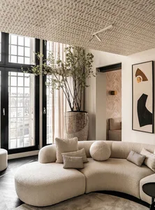 Modern minimalist living room with a plush, neutral-colored sectional sofa, large potted plant, and a patterned wallpaper cei