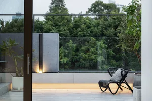 Black wicker chair with white cushion on a concrete patio bordered by glass railings, surrounded by lush greenery and trees.
