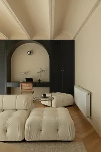 A minimalist living room with a white fabric sofa, ottoman, and a black desk with a chair, all set against a cream-colored wa