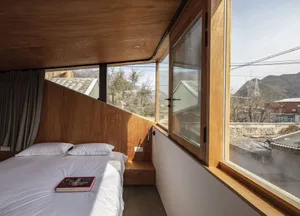 A modern bedroom with a bed, pillows, and a book on a wooden floor, large windows, and a view of the mountains.