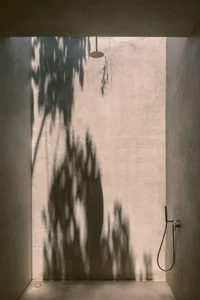 A minimalist concrete bathroom features a circular shower head and a shadow of trees cast on the wall, creating a serene and 