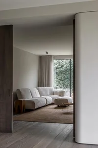 A minimalist living room with a beige fabric sofa, a wooden coffee table, and a large window overlooking greenery.