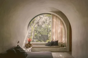 Modern bedroom featuring a concrete sitting area with natural light from a large arched window overlooking trees.