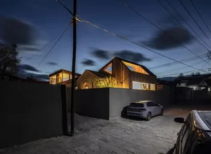 A modern wooden house with illuminated windows at dusk, surrounded by a concrete wall and a parked car.