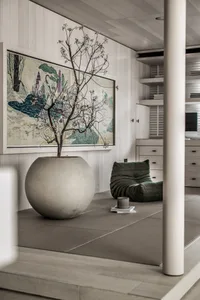 A serene living room with wooden floors, a large decorative pot with plants, and a minimalist green seat.