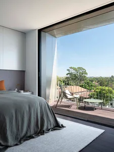 A modern bedroom with a wooden floor, featuring a bed, a rug, and a large window overlooking a balcony with a chair and table