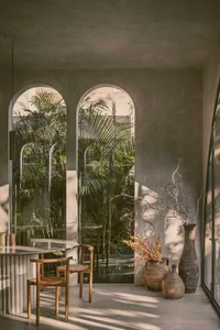 A modern, minimalist living room with concrete walls and floor, featuring a clear view of palm trees outside through arched w