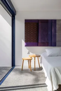 A modern bedroom featuring a bed with gray bedding, wooden stools, and a wall art piece with purple and brown panels.