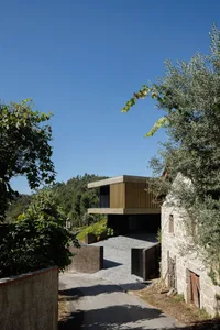 A modern house with a concrete and wood exterior sits nestled among green trees with a gravel driveway leading up to it.