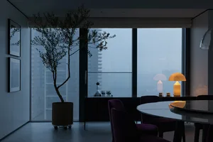 A modern dining room with a round table, purple chairs, and a contemporary lamp. Large windows offer a cityscape view.