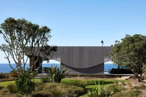 A modern house with a curved roof and concrete exterior walls sits on the coast.