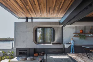 Modern residential kitchen featuring concrete walls and a curved window with horizontal black bars, overlooking a serene wate