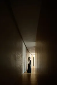 A woman in a black dress standing in a dark hallway with wooden walls and a gray light on the end.