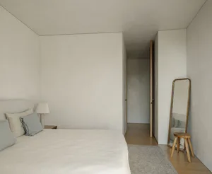 A minimalist bedroom with a bed, lamp, mirror, stool, and rug.