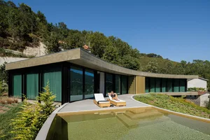 A modern holiday villa featuring a concrete and glass exterior with a swimming pool and lounge chairs.