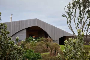 A modern house with a curved metal roof and greenery surrounding it.
