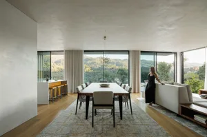 A modern minimalist dining room with a large table, chairs, and a view of the mountains through floor-to-ceiling windows.