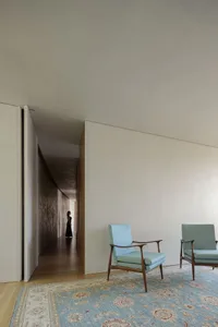 A minimalist living room with fabric-covered walls and wooden furniture, featuring a blue armchair on a floral-patterned rug.