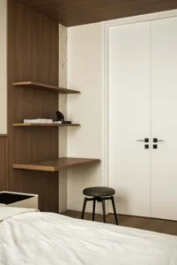 A minimalist bedroom with a white bed, wooden shelves, marble accent wall, and black stool.