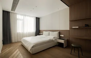 A minimalist bedroom with a white bed, wooden paneling, white curtains, and a window letting in natural light.