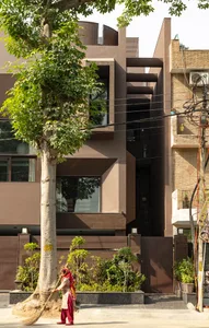 A modern residential building with glass and brick exterior materials, featuring a tree and a woman sweeping the sidewalk.