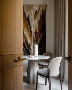 A modern dining room featuring a round marble table with white chairs, a vase with dried flowers, and a textured abstract art