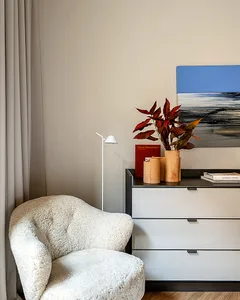 Modern living room with a white sheepskin upholstered chair, concrete floor, and a bookshelf with a plant and books.