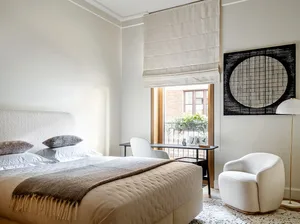 A minimalist bedroom featuring white furniture, a fabric-covered bed, and a window with natural light.