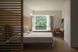 A minimalist bedroom featuring a bed with white sheets and a wooden headboard and floor, with a large window showcasing a lus
