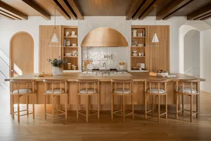 A modern residential kitchen with wooden cabinets and flooring, featuring a large island, pendant lights, and a stove with a 