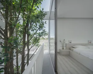 A minimalist bedroom with a white bed and wooden floor, featuring green trees outside the large windows and a clean, modern d