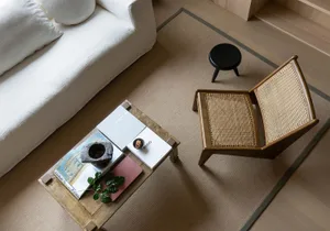 A living room with white sofa, woven armchair, coffee table, and black stool on a beige rug.