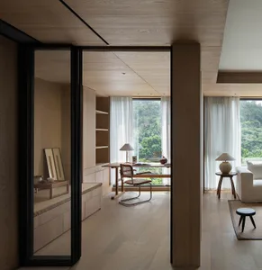 A minimalist wooden room with a desk, chair, and large windows overlooking greenery.