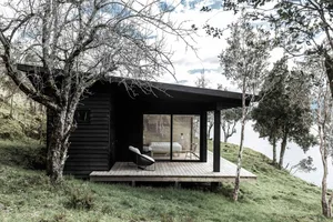 A minimalist black cabin with a wooden deck and glass windows, situated by a body of water.