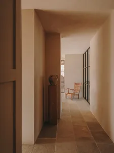 A minimalist corridor featuring stone flooring and walls with a vintage vase on a pedestal and a woven chair.