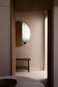 A minimalist bedroom featuring a circular mirror on the wall, a wooden bench, and sheer curtains, creating a serene and moder