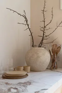 A minimalist interior design space featuring a ceramic vase with dried branches and a stack of ceramic dishes on a marble cou