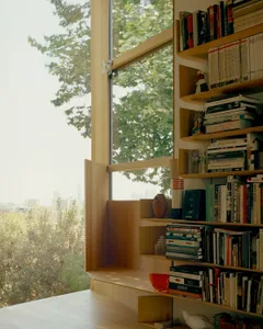 A modern bookshelf filled with books, vases, and a speaker, next to a large window with a view of trees.