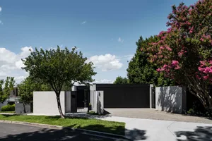 A modern house with a black gate and concrete walls, surrounded by flowering trees on a clear day.