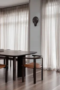 A modern dining room featuring wooden furniture, leather seating, light-colored sheer curtains, and wall-mounted light fixtur