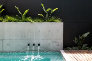 A modern swimming pool with water spouts and a concrete wall, featuring green plants and a wooden deck.