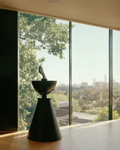 A modern sculpture with a cone-shaped base and a bowl-like structure on top, set against a large window with a view of a city