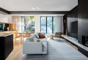 A modern living room with a grey sofa on a grey rug, wooden flooring, a kitchen counter with vases, and large windows overloo